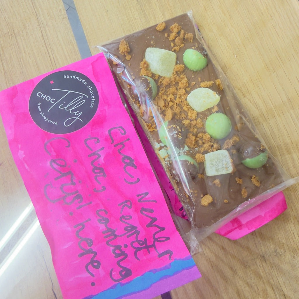Chocolate bar with green candy pieces in clear packaging on a pink bag with text.